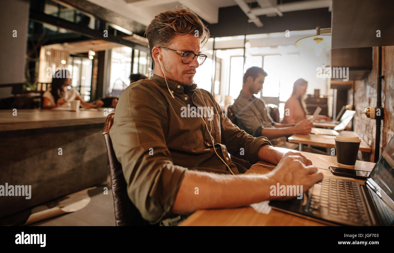 Junger Mann im Café sitzen und mit Laptop-Computer, mit Menschen im Hintergrund. Stockfoto