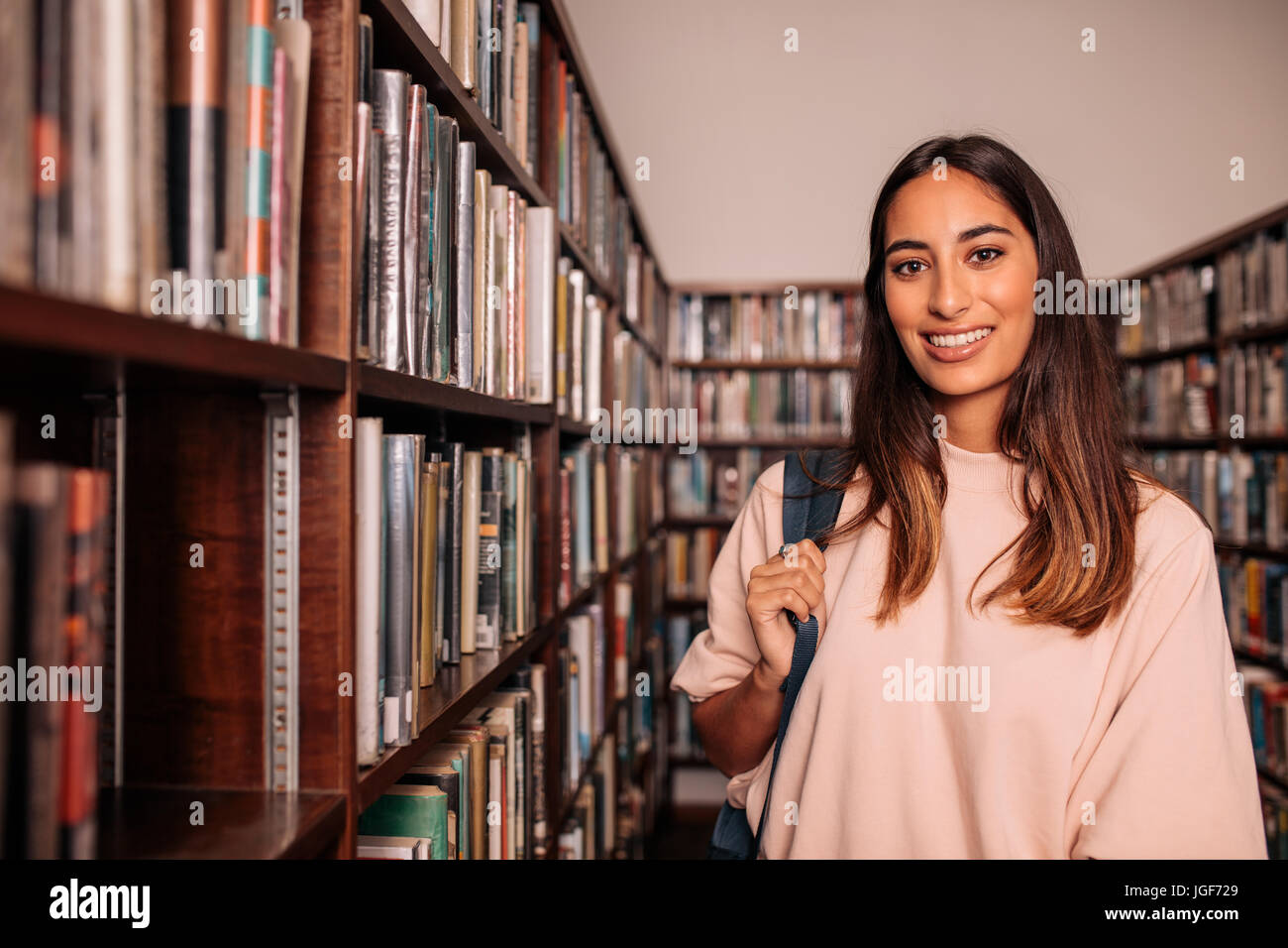 Junge Studentin in der Bibliothek stehen. Porträt der glückliche junge Frau mit Tasche stehen in der öffentlichen Bibliothek von den Bücherregalen. Stockfoto