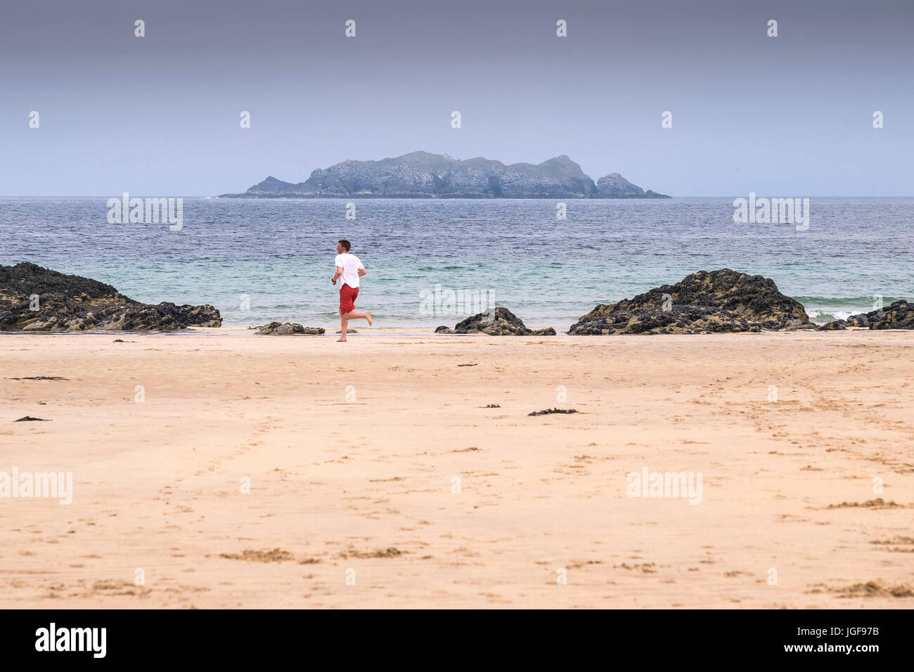 Ein Mann, der am Strand von Harlyn Bay mit Gulland Island im Hintergrund läuft. North Cornwall. Stockfoto