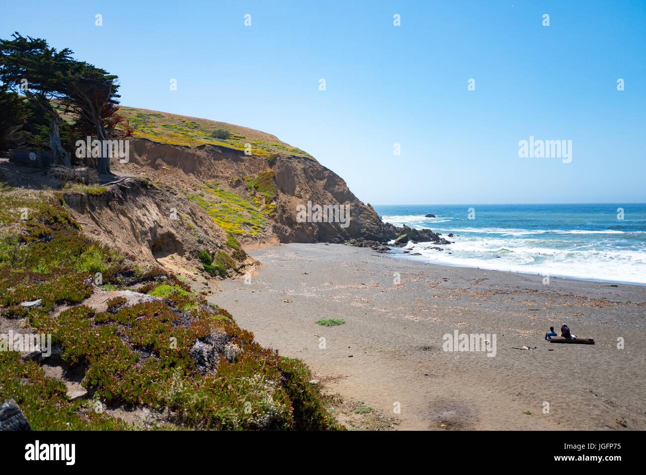 Zwei Menschen sitzen auf einem Baumstamm an einem einsamen Strand mit Wellen, die an der Küste, Mori Point, Teil der Golden Gate National Recreation Area in Pacifica, Kalifornien, 20. Juni 2017. Stockfoto