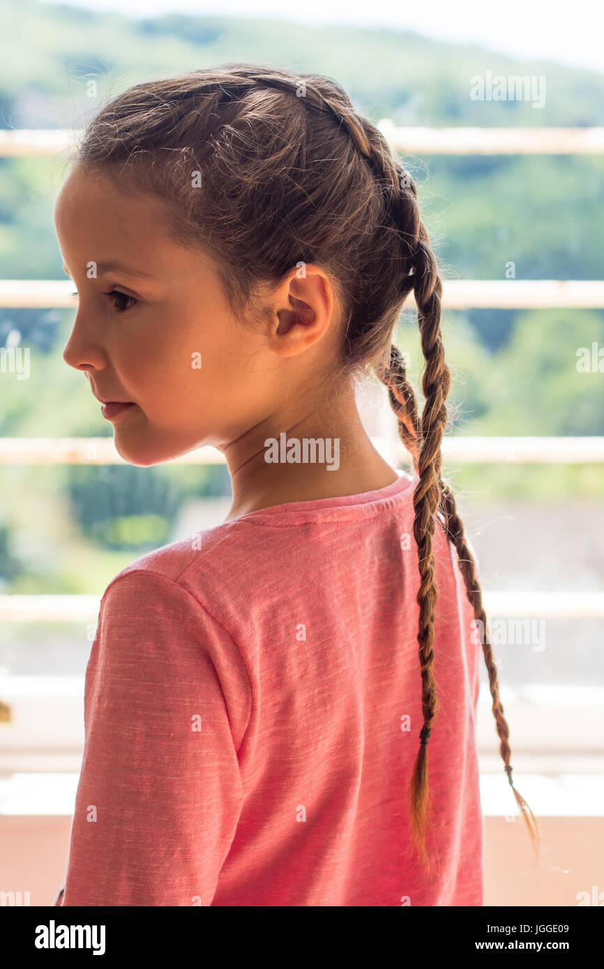 Mädchen mit niederländischen Zöpfen Blick aus Fenster im Profil. Kleines Kind mit braunen Haaren geflochten in einem rosa top Stockfoto