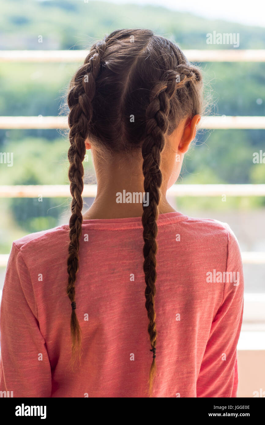 Mädchen mit niederländischen Zöpfen Blick aus Fenster von hinten. Kleines Kind mit braunen Haaren geflochten in einem rosa top Stockfoto