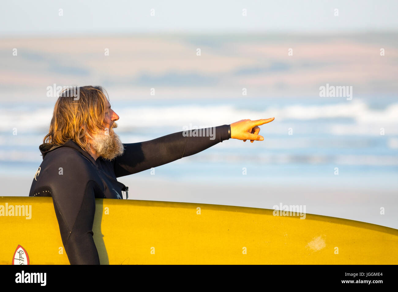 Ein reifer männlicher Surfer mit langem Bart hält ein Surfbrett und deutete in Richtung Meer auf der beliebten Surf Strand Polzeath, Cornwall Stockfoto Ein reifer männlicher Surfer mit langem Bart hält ein Surfbrett und deutete in Richtung Meer auf der beliebten Surf Strand Polzeath, Cornwall Stockfoto