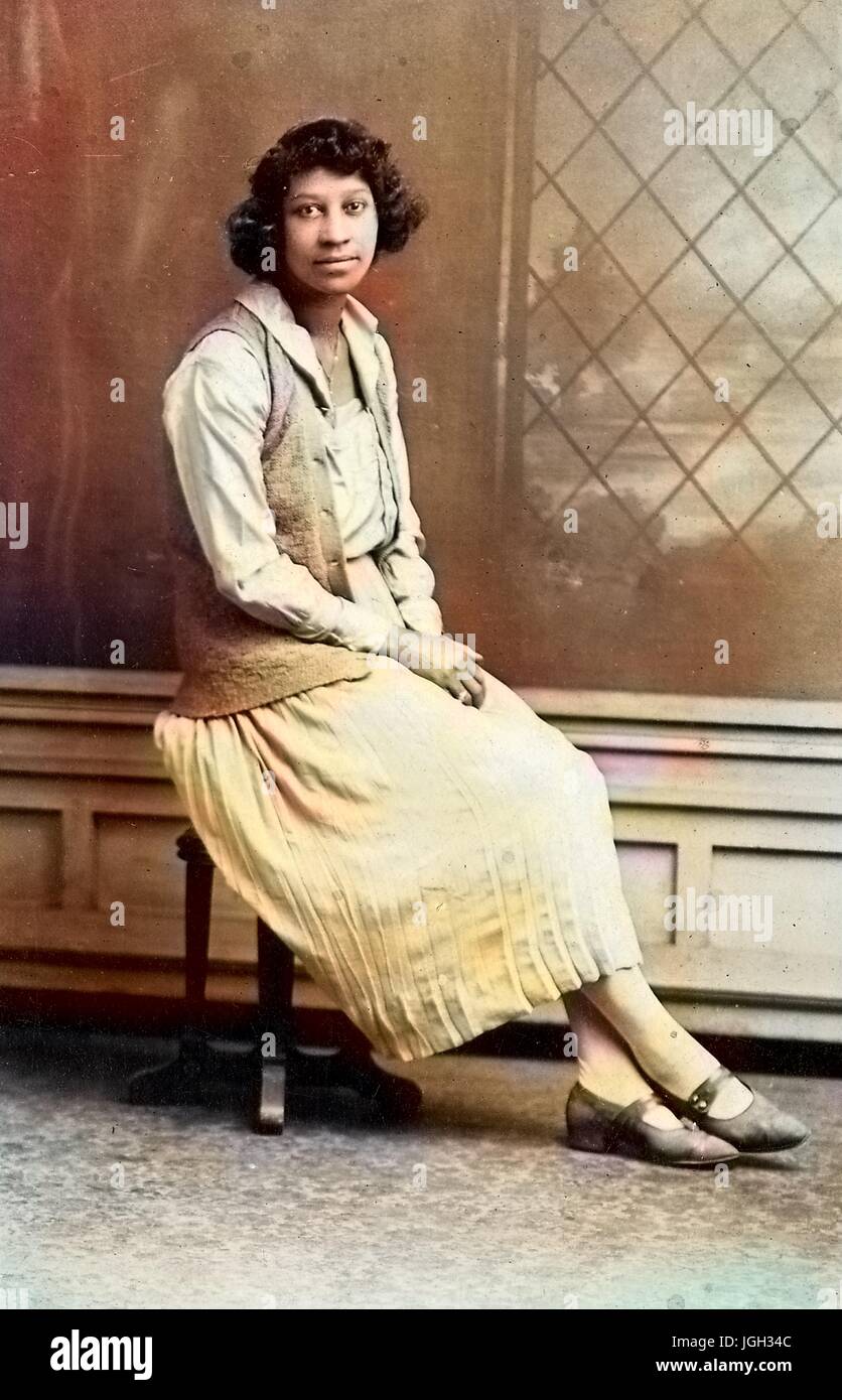 Voller Länge sitzen Portrait, junge afroamerikanische Frau, trägt leichte Kleid und Weste, sitzen auf Stuhl vor Kulisse, neutralen Gesichtsausdruck, 1920. Hinweis: Bild wurde digital eingefärbt wurde mit einem modernen Verfahren. Farben können Zeit ungenau sein. Stockfoto