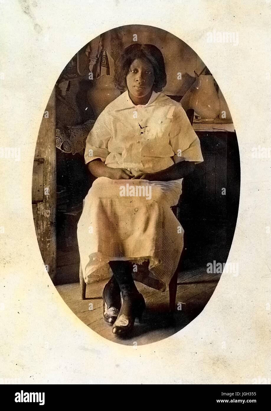 Voller Länge sitzen Portrait, Reife afroamerikanische Frau, leichtes Kleid, sitzt vor Schrank mit Vasen, neutralen Gesichtsausdruck, 1915. Hinweis: Bild wurde digital eingefärbt wurde mit einem modernen Verfahren. Farben können Zeit ungenau sein. Stockfoto