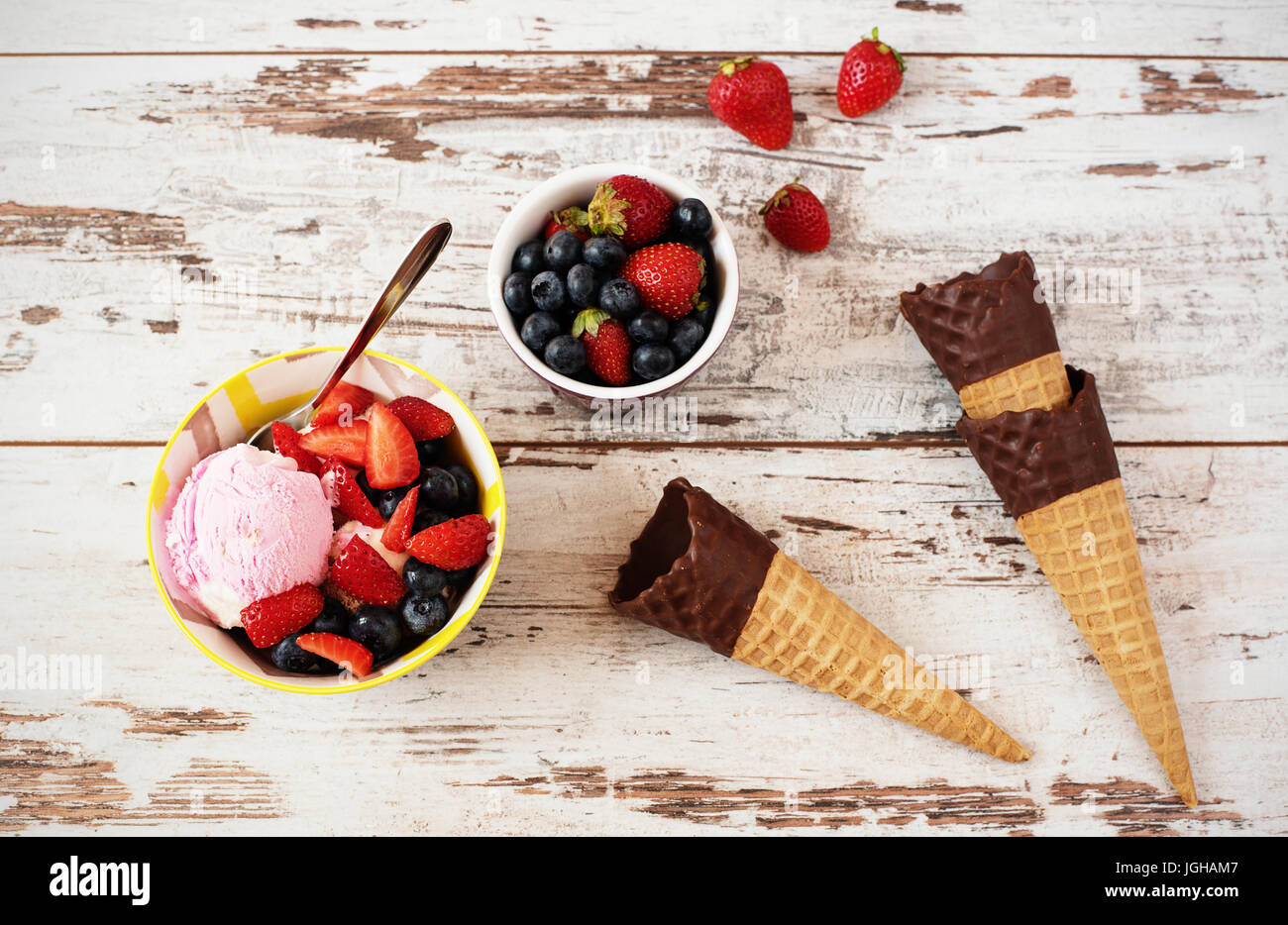 Rosa Eis serviert mit Beeren - Erdbeeren und Heidelbeeren in eine gelbe Schale. Waffelkegeln mit Schokolade. Hellen rustikalen hölzernen Hintergrund Stockfoto