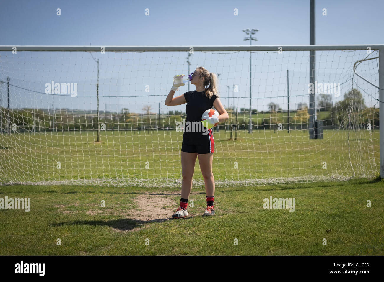 Gesamte Länge der weibliche Fußball Spieler Trinkwasser gegen Torpfosten auf Feld Stockfoto
