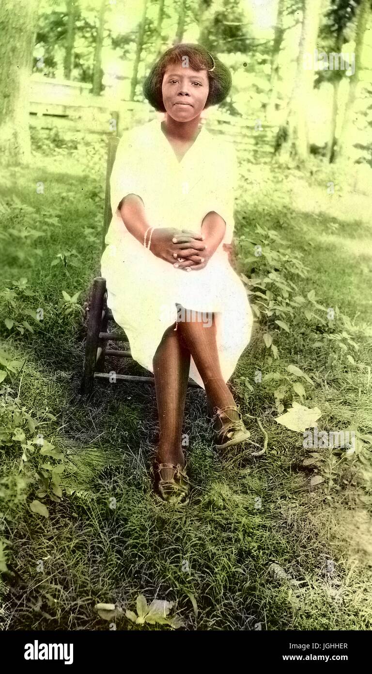 Young African American Frau posieren für ein Porträt in einem bewaldeten clearing, Schneidersitz auf einem Stuhl sitzend und trägt ein weißes Kleid mit einer ernsten Gesichtsausdruck, 1944. Hinweis: Bild wurde digital eingefärbt wurde mit einem modernen Verfahren. Farben können Zeit ungenau sein. Stockfoto