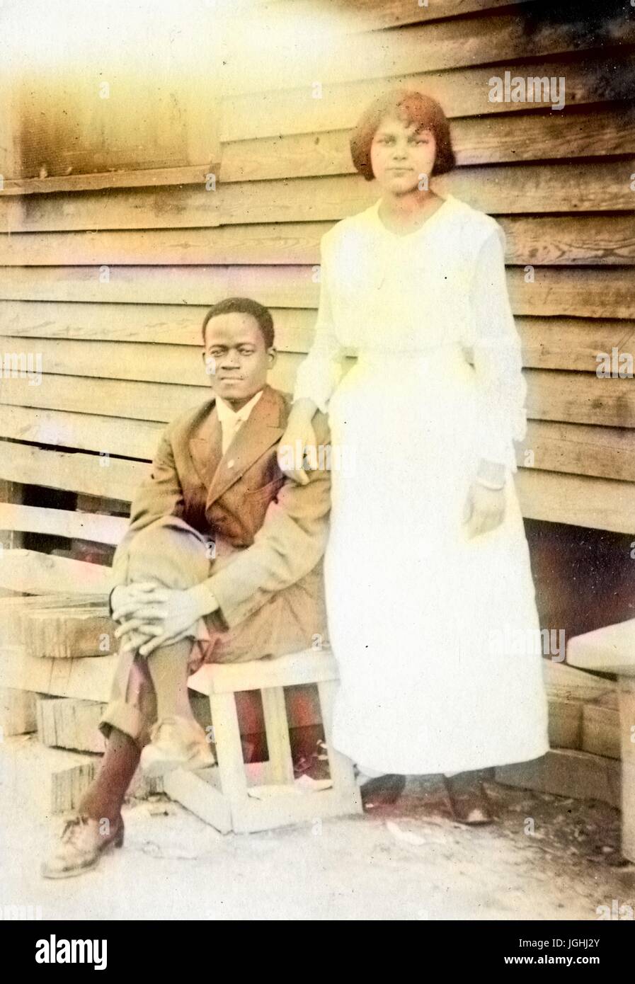 African-American Paar posieren vor einem Haus mit Holz Abstellgleis, Frau trägt weißes Kleid und stehend, Mann sitzt auf einer Holzkiste mit seiner Hand auf sein Knie, 1910. Hinweis: Bild wurde digital eingefärbt wurde mit einem modernen Verfahren. Farben können Zeit ungenau sein. Stockfoto