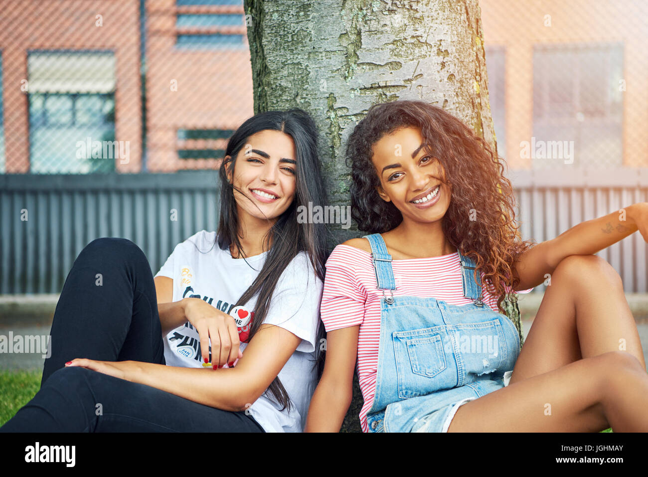 Zwei attraktive junge Freunde sitzen zusammen auf einer Wiese schiefen Lächeln zurück auf einem Baumstamm an einem sonnigen Tag Stockfoto