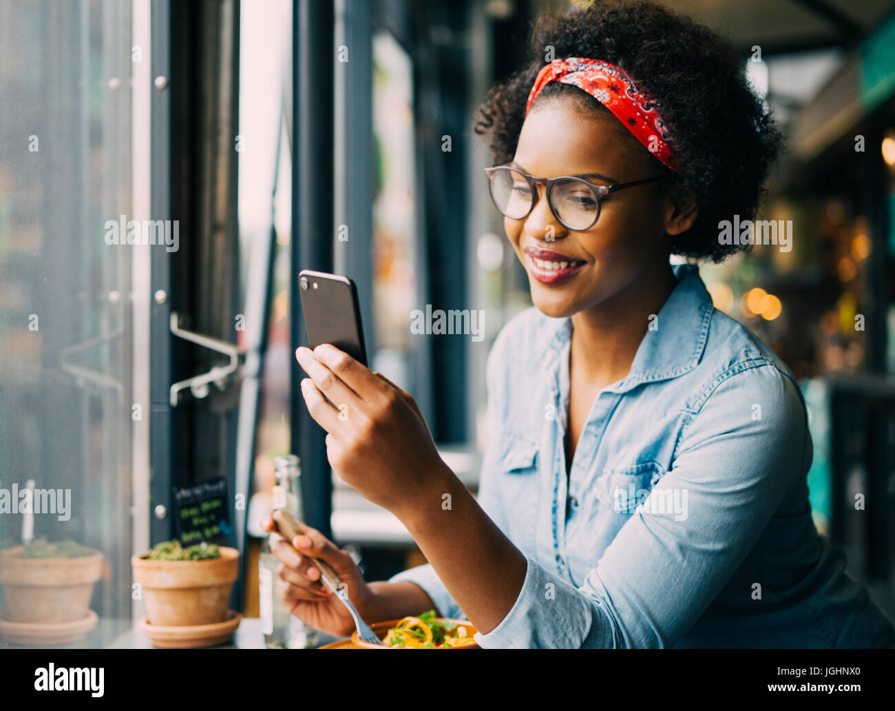 Attraktive junge afrikanische Frau lächelnd und Lesen von Texten auf ein Handy beim Sitzen allein an einem Schalter in einem Café genießen Sie eine Mahlzeit Stockfoto