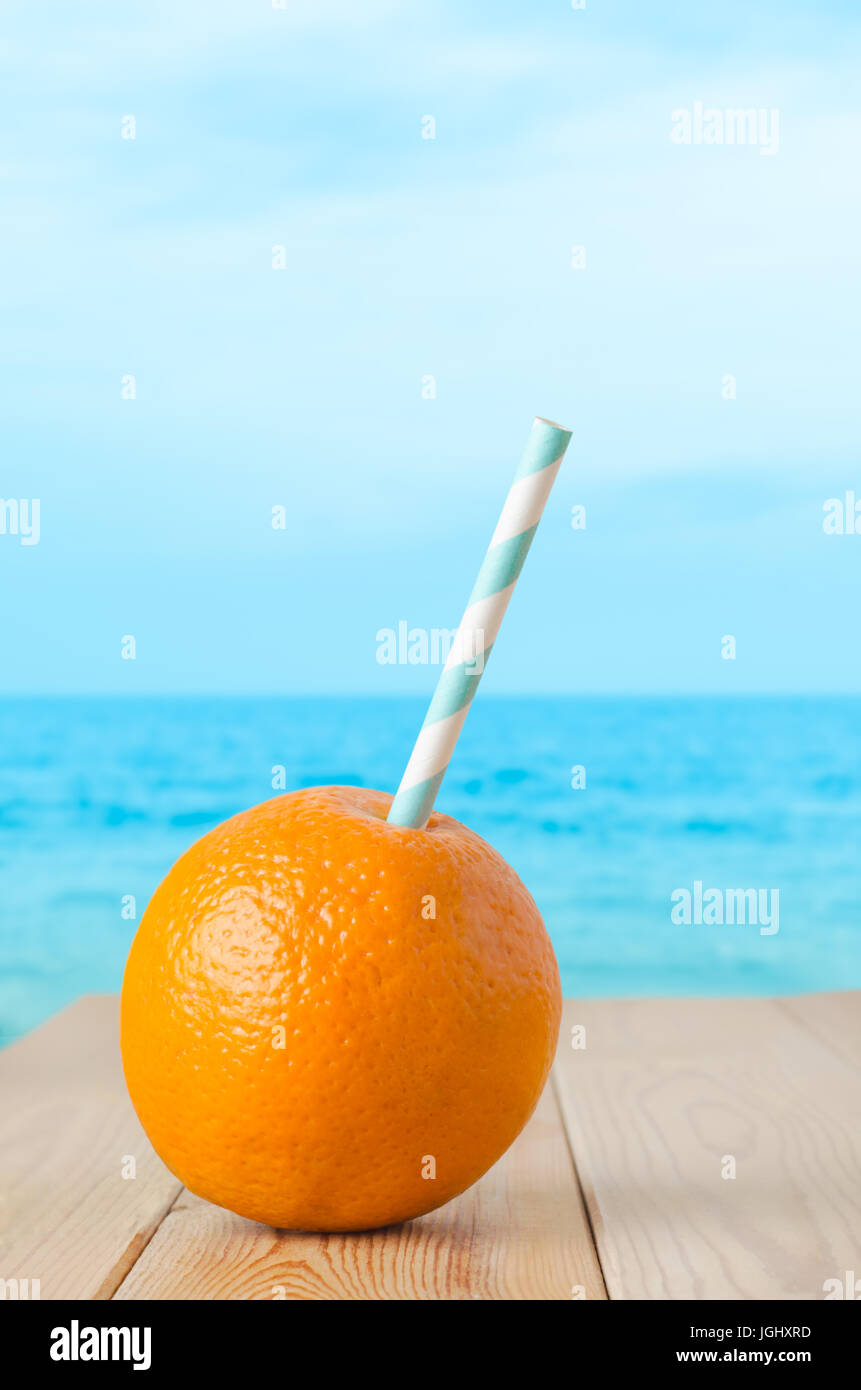 Eine ganze Orange, durchbohrt mit einem gestreiften Strohhalm, ein frisches Obst-Getränk zu vertreten.  Stellen Sie auf einem geplankte Holztisch mit Meer und Himmel Hintergrund weichzeichnen. Stockfoto
