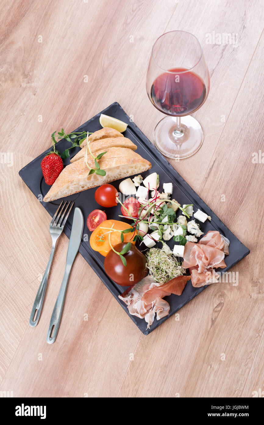 Snacks mit Rotwein auf schwarzem Schiefer an Bord über hölzerne Hintergrund. Delikatesse und einfaches Essen, Brot, Käse, Tomaten und Schinken. Stockfoto