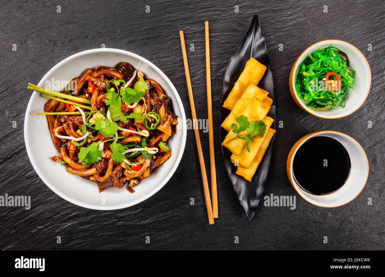 Asiatische Nudeln mit würziger Sojasauce, Hähnchenteile und Frühlingsrollen, Draufsicht, serviert auf schwarzem Stein. Stockfoto