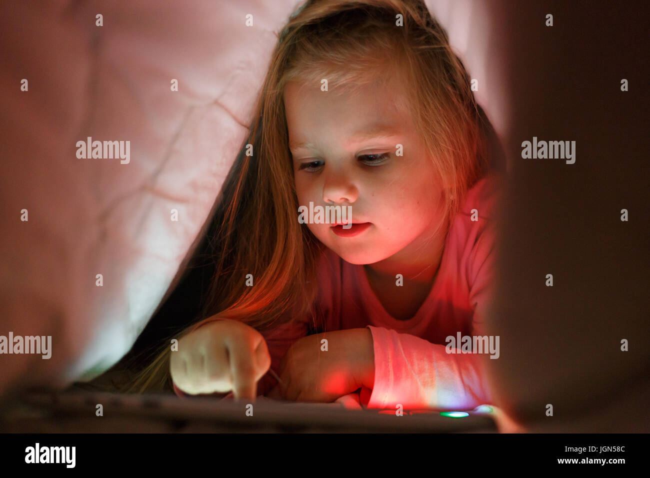 Kleines Mädchen spielt auf dem Tablet im geheimen von ihren Eltern in der Nacht unter der Decke Stockfoto