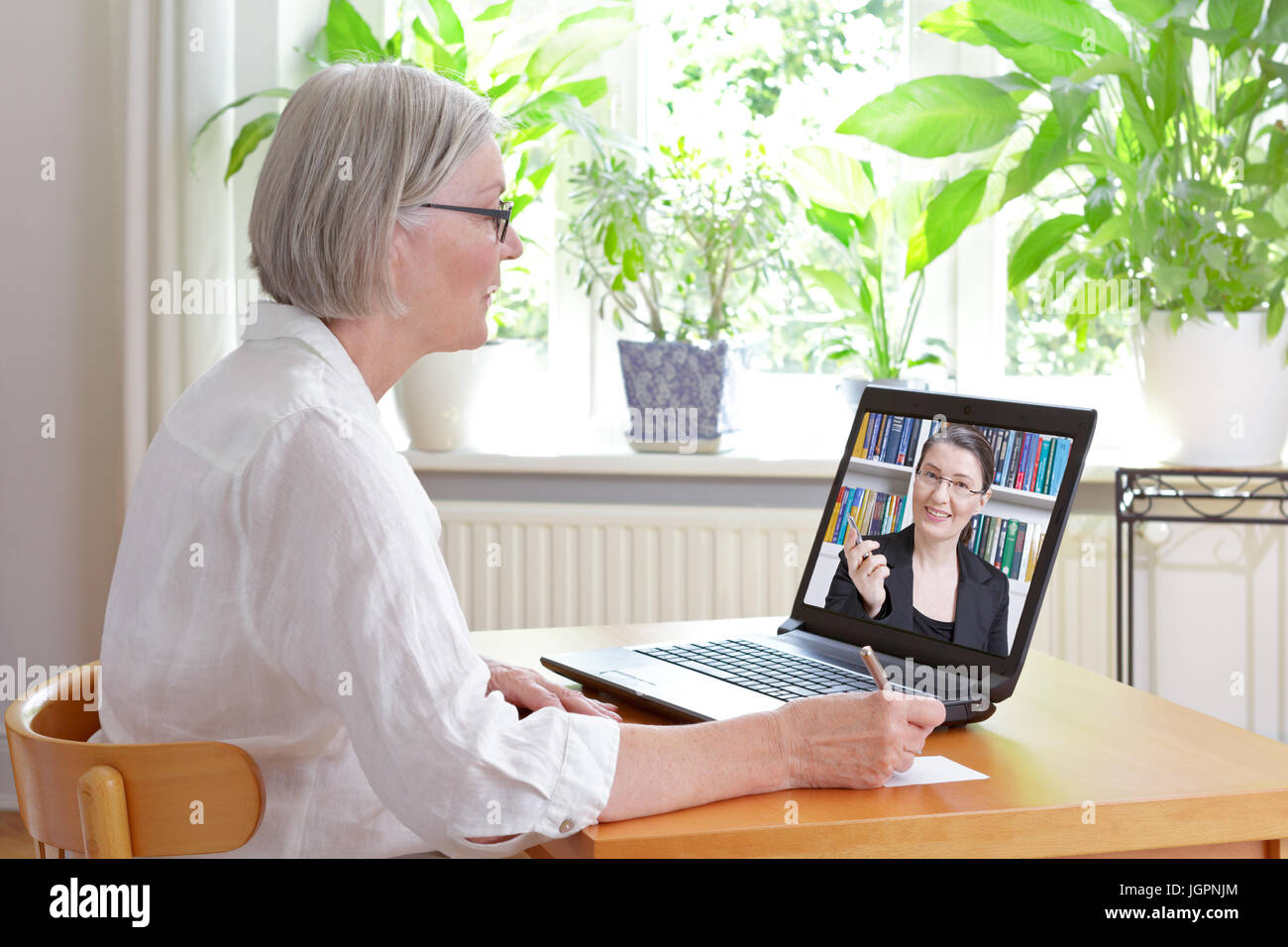 Ältere Frau zu Hause vor ihrem Laptop Notizen während gerade eine Online-Video von Steuerberatung durch einen weiblichen Buchhalter Stockfoto