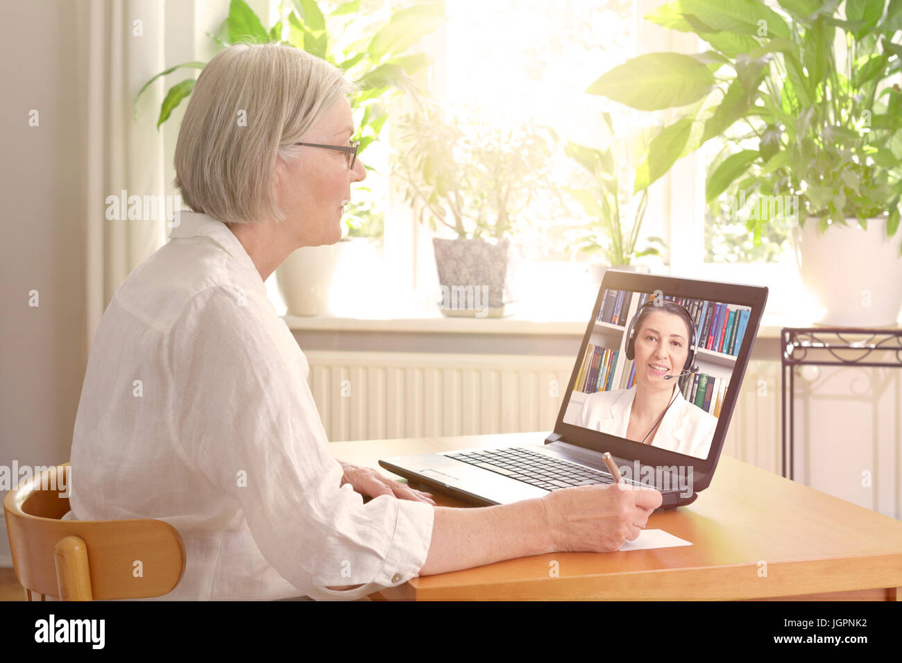 Ältere Frau in ihrem sonnigen Wohnzimmer vor einem Laptop Notizen während gerade eine Online-Beratung durch einen weiblichen Therapeuten video Stockfoto