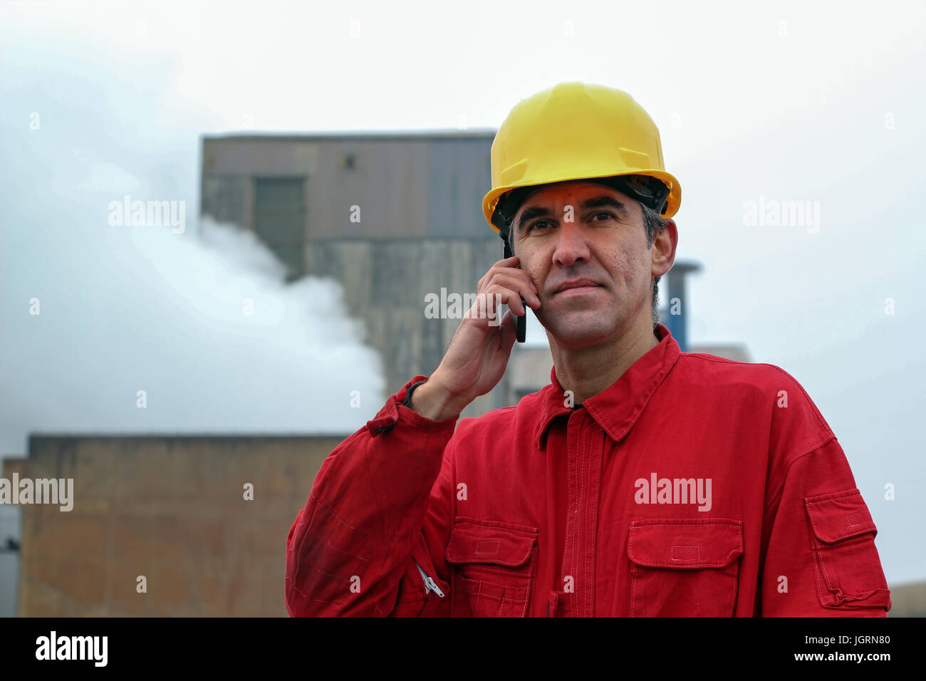 Ein Portrait eines Arbeitnehmers im roten Overall und gelben Helm mit Handy vor dem Schornstein Rauchen. Stockfoto