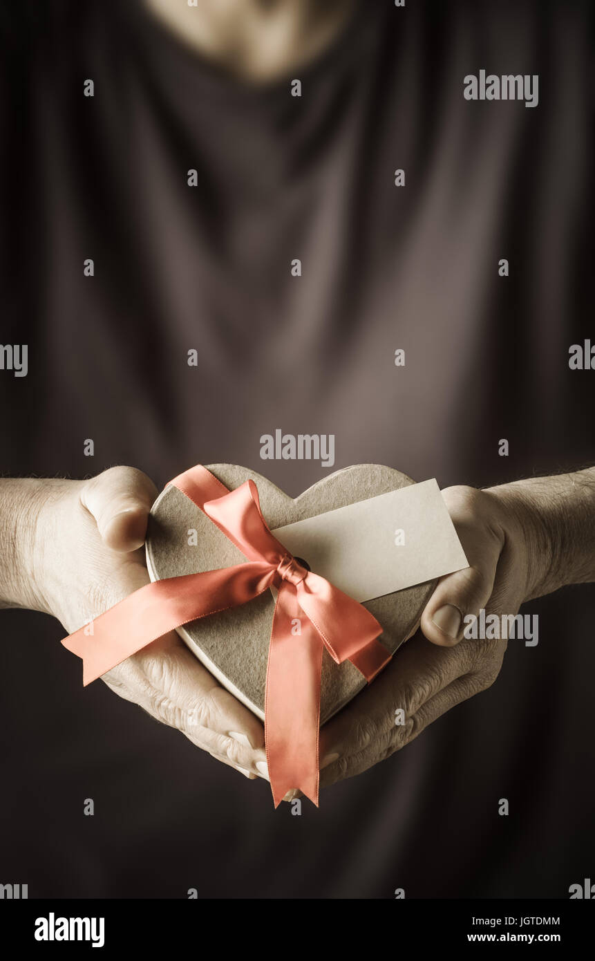 Männliche Hände halten ein Herz geformt Geschenk-Box, um einen Bogen mit Satinband gebunden.  Eine leere Geschenk Label zeigt Frontseite um Nachricht textfreiraum vorsehen.  M Stockfoto