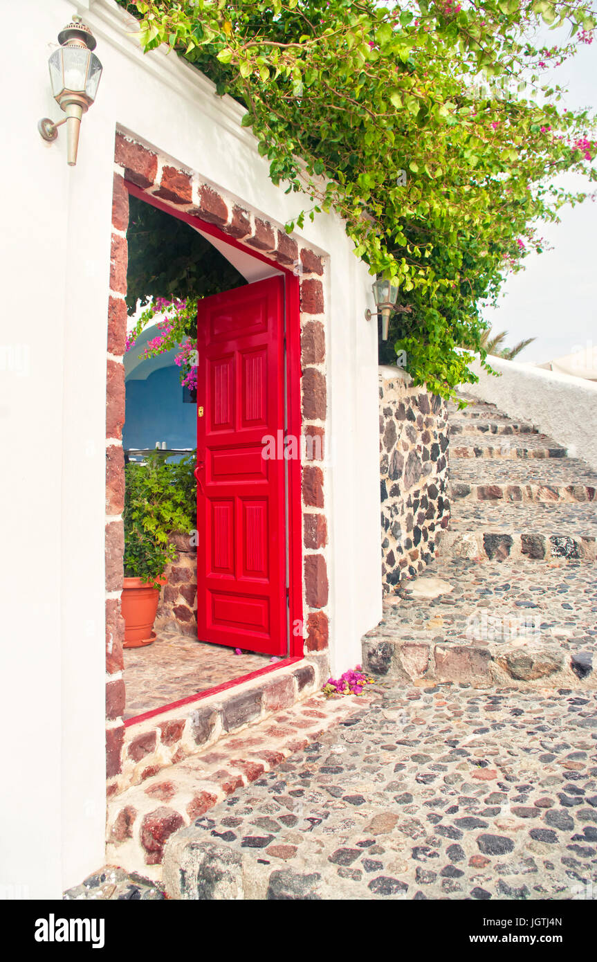 Rote offene Tür eines Hauses mit blühenden Pflanze drüber an warmen Frühlingstag mit Boulder Treppen in Oia, Santorini oder Thira, Griechenland Stockfoto