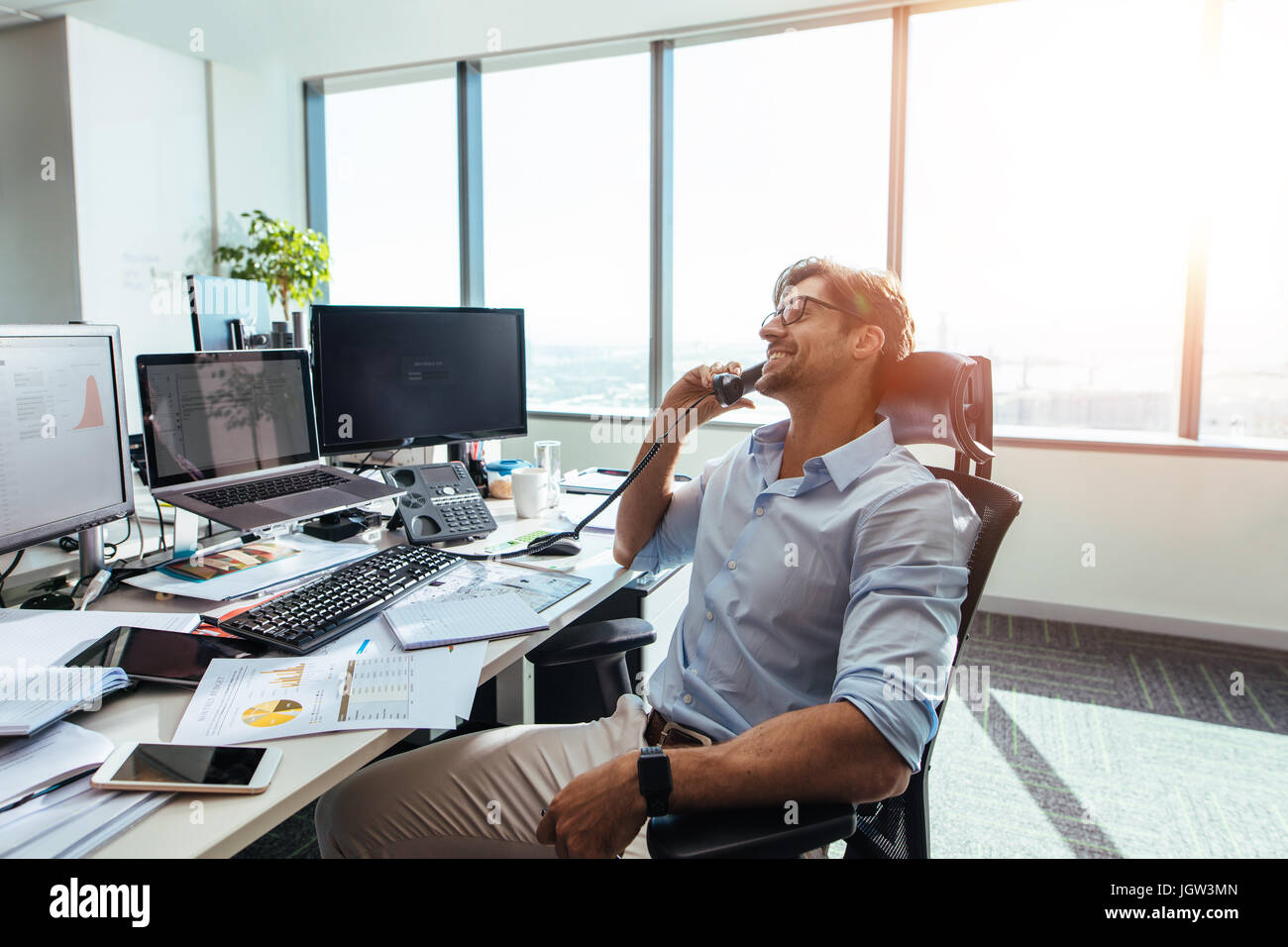 Glückliche Unternehmer an seinem Schreibtisch im Büro sprechen über Telefon. Junger Geschäftsmann im Büro mit Computer und Geschäftspapiere am Tisch sitzen. Stockfoto