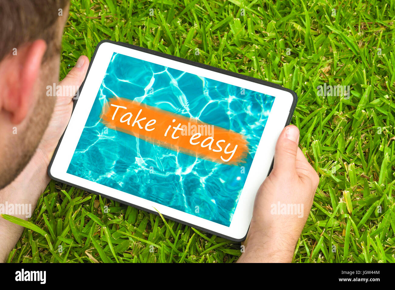 Junger Mann liegend auf dem Rasen im Garten mit Blick auf Wasser Hintergrund mit Satz "Take it easy" darauf geschrieben Tablet-Gerät. Stockfoto