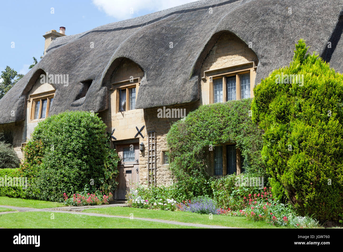 Traditionelles Steinhaus mit Strohdach und Blumen, Sträucher im Vorgarten, in einem englischen Dorf in Cotswolds, an einem sonnigen Sommertag. Stockfoto