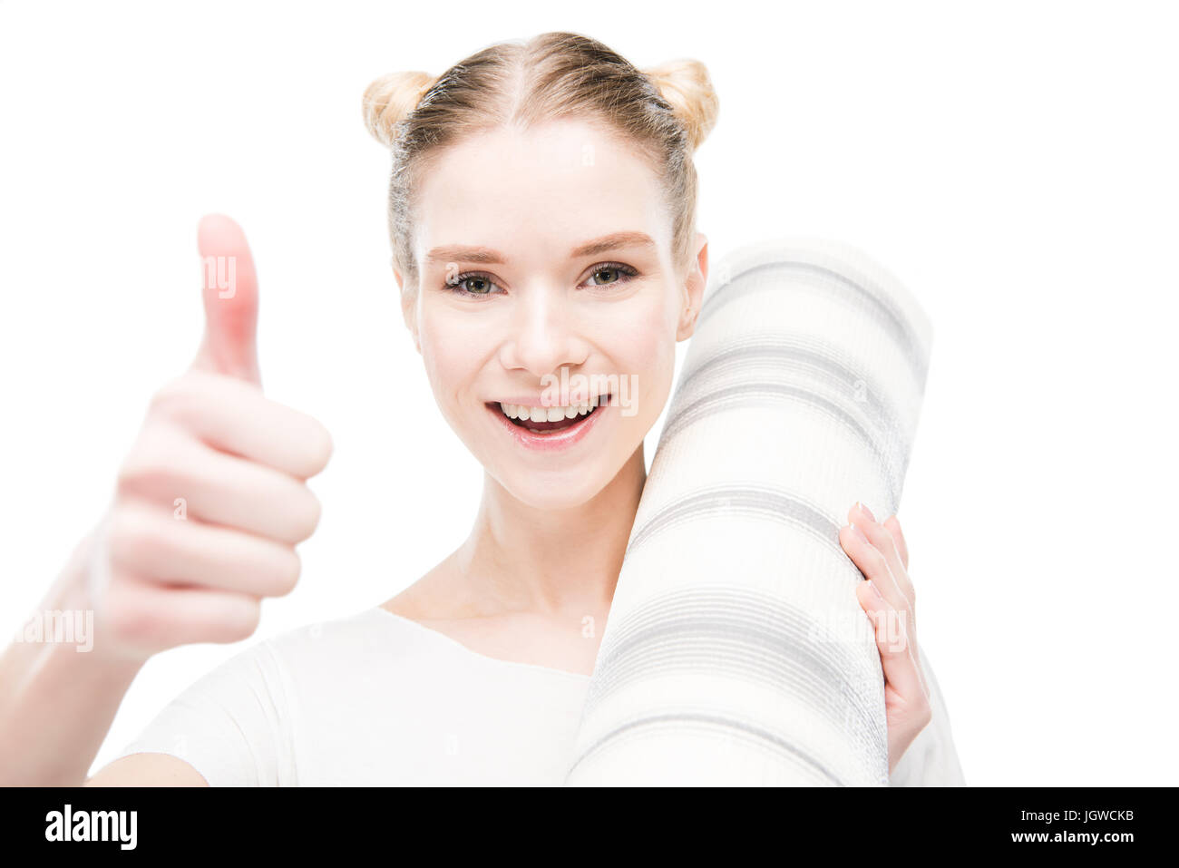 Lächelnde junge Frau hält Yoga-Matte und zeigt Daumen oben isoliert auf weiß Stockfoto