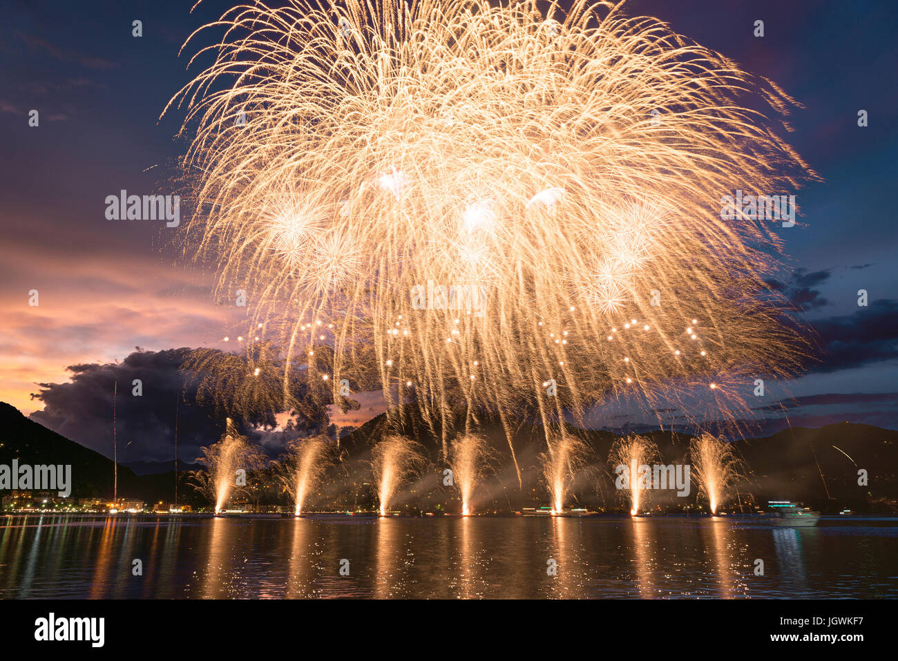 Feuerwerk auf dem Luganer See an einem Sommerabend mit bewölktem Himmel bei Sonnenuntergang im Hintergrund Stockfoto