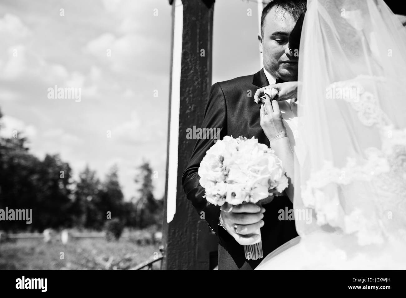 Wunderschöne junge Braut pinning Knopfloch-Blume des Bräutigams Jacke outdoor am Tag ihrer Hochzeit. Schwarz / weiß Foto. Stockfoto