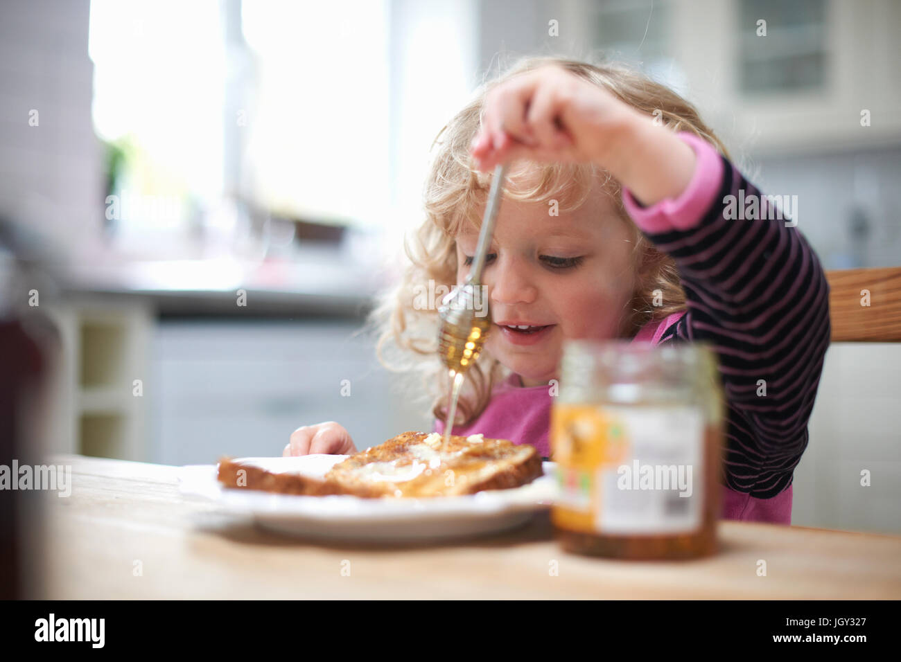 Junges Mädchen saß am Küchentisch, nieselte Honig auf toast Stockfoto