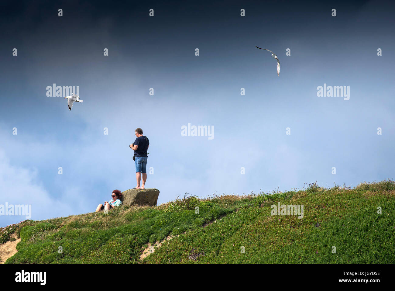 Ein Mann steht auf einem Felsen mit seiner Kamera und eine Frau zum Entspannen in der Sonne auf der Landzunge wie Möwen über Kopf fliegen.  Newquay, Cornwall. Stockfoto