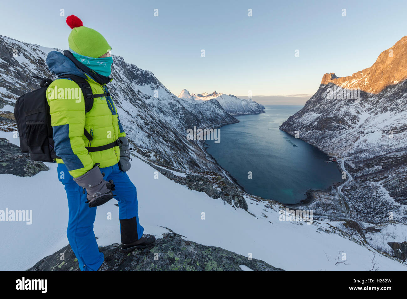 Wanderer auf dem Kamm bewundert das Dorf Fjordgard umrahmt das gefrorene Meer des Ornfjorden, Senja, Troms, Norwegen Stockfoto