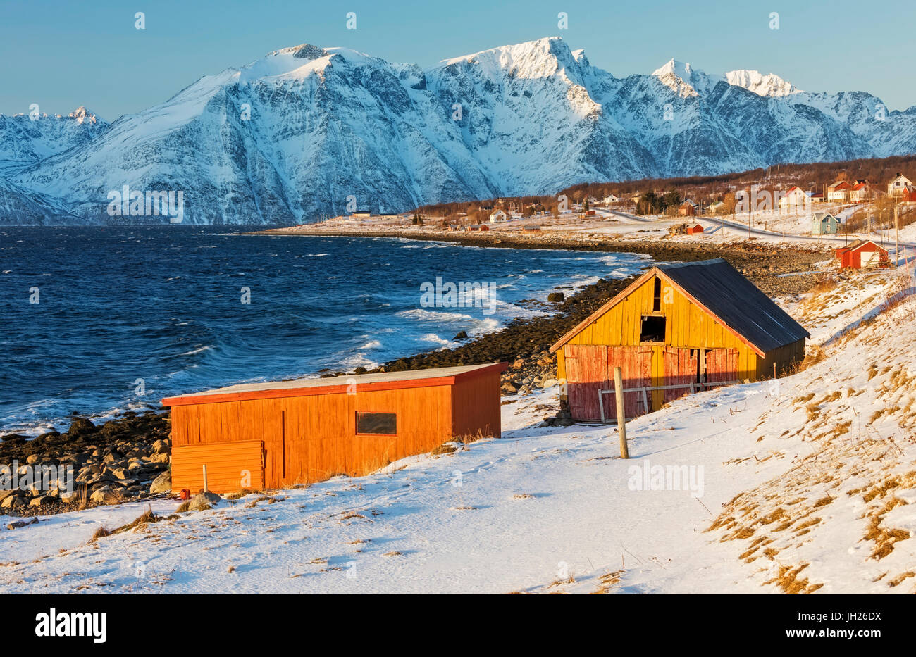 Typische Holzhütten genannt Fischerorten umgeben von Wellen der kalten See und schneebedeckten Gipfeln, Djupvik, Lyngen Alpen, Troms, Norwegen Stockfoto