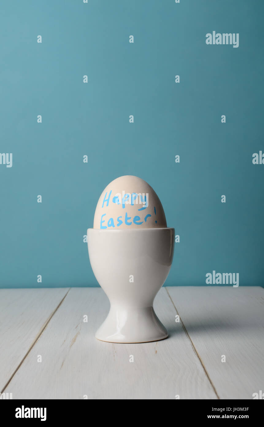 Ein künstlicher Ei in einen Eierbecher mit Worten "Frohe Ostern" in blau gemalt.  Küchentisch weiß Plank und blauem Hintergrund. Stockfoto
