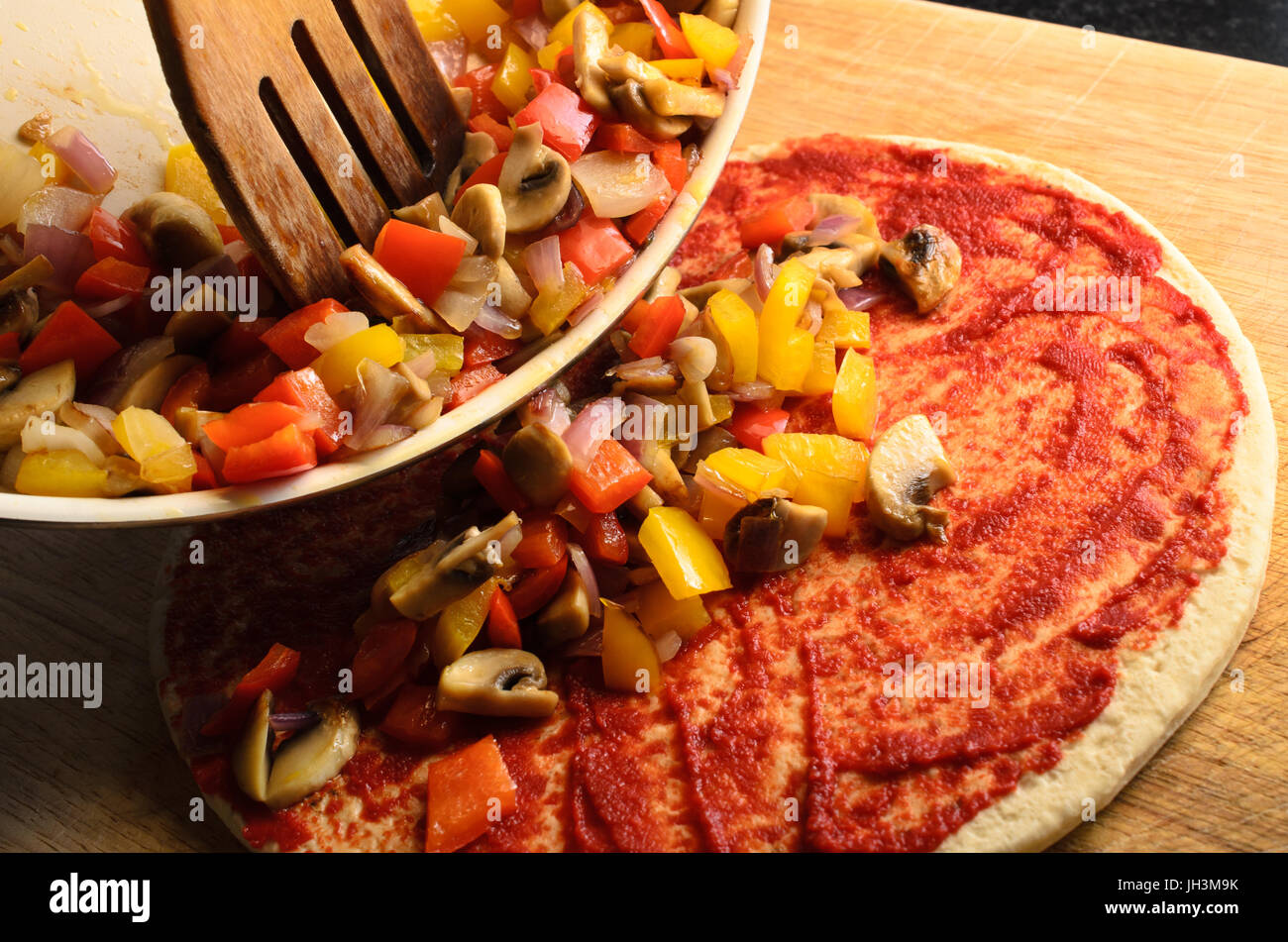 Pizza Vorbereitung Szene.  Gemischtes, gebratenes Gemüse auf Pizza aus der Pfanne mit Holzspatel gekippt wird. Stockfoto