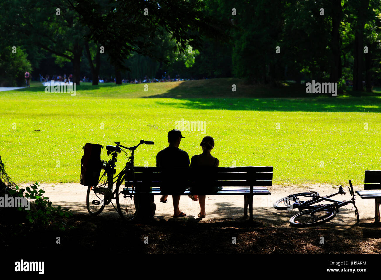 Hintergrundbeleuchtung junges Paar mit Fahrräder sitzen auf einer Parkbank, Englischer Garten, München, Bayern, Deutschland Stockfoto