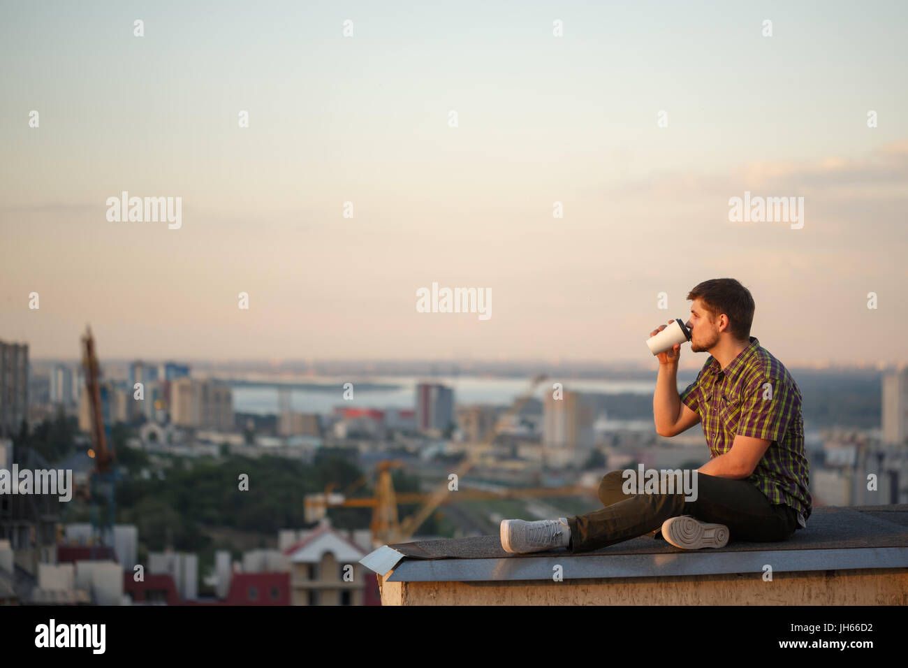 Ein Mann trinkt Kaffee in den frühen Morgenstunden auf dem Dach. Er schaut Weg und denkt. Im Hintergrund die Stadtlandschaft Stockfoto