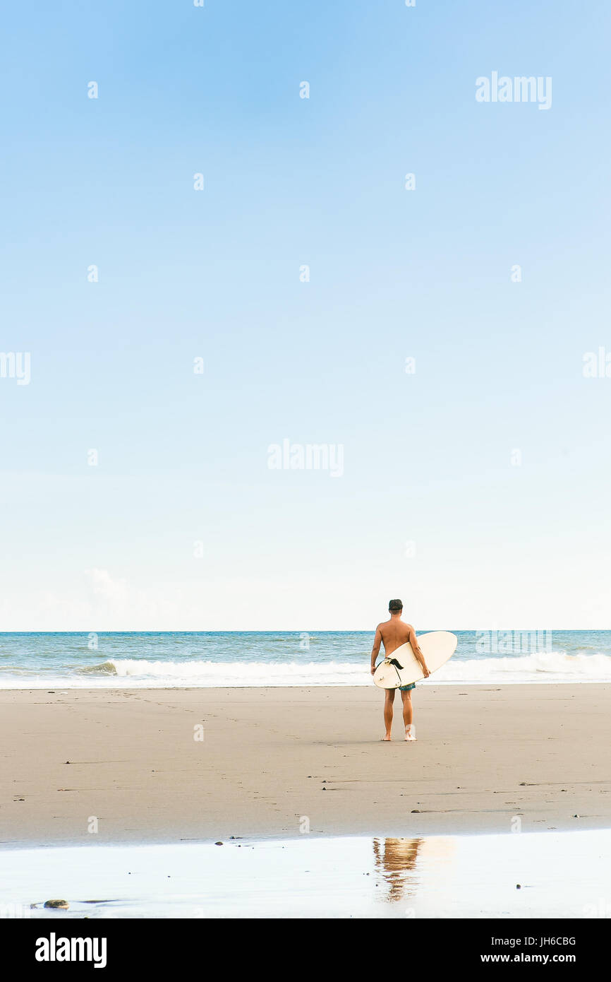Hübscher Junge in blau Badeshorts und Mütze mit langen Surf Board warten auf Surfspot am Meer Ozeanstrand Surfen stehen. Weiße leere Surfbrett. Konzept-o Stockfoto