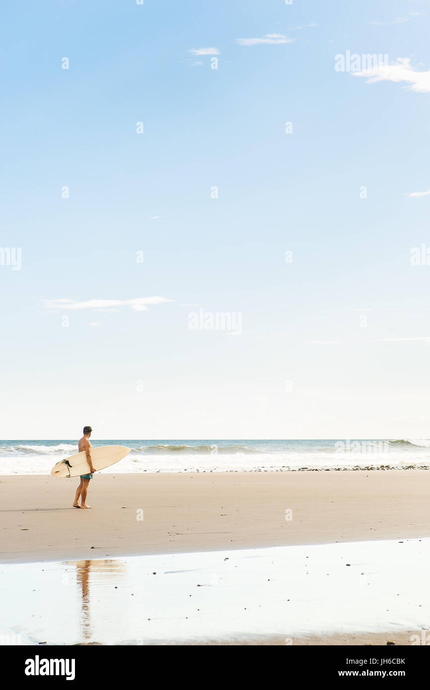 Hübscher Junge im blauen Badeshorts und Kappe gehen mit lange Surf Surf Board warten auf Surfspot am Meer Ozeanstrand. Weiße leere Surfbrett. Konzept der Stockfoto