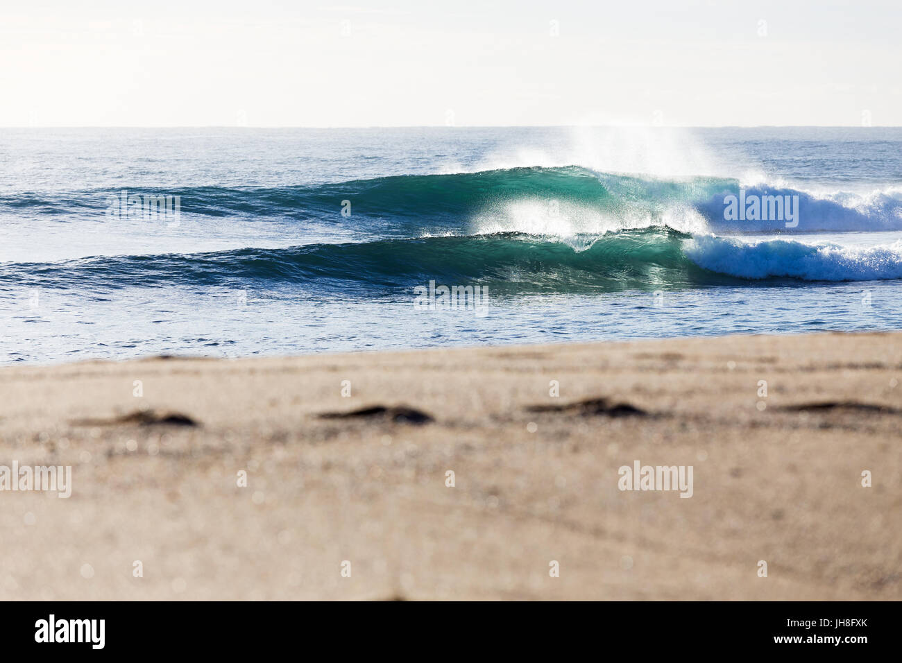 Frische Meeresluft Wellen brechen im hellen Morgenlicht zu einem schönen Sandstrand mit Fußspuren im Vordergrund. Stockfoto