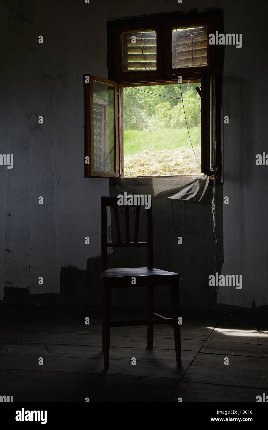 Stuhl vor einem offenen Fenster mit einem Moskitonetz. Eine dunkle indoor-Einstellung: schäbig Wand, rustikales Holz Planke Bodenbelag. Stockfoto