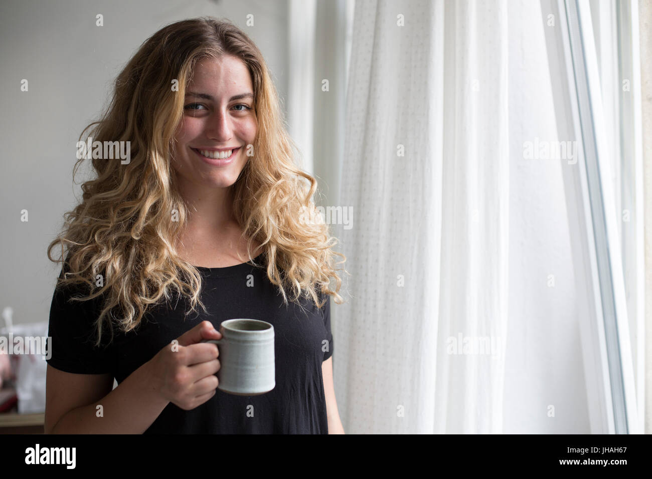 Jung, blond, schöne kaukasischen Frau bei einem heißen Getränk in der Nähe der Fenster im natürlichen Morgenlicht. Drinnen im Sommer auf einer Canon 5D aufgenommen Stockfoto