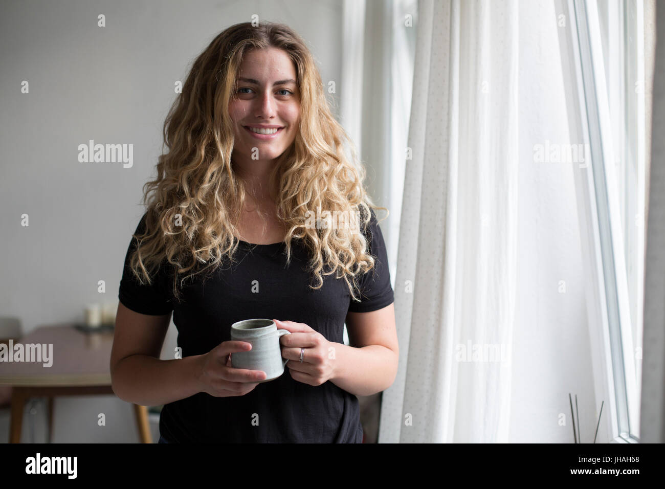 Jung, blond, schöne kaukasischen Frau bei einem heißen Getränk in der Nähe der Fenster im natürlichen Morgenlicht. Drinnen im Sommer auf einer Canon 5D aufgenommen Stockfoto
