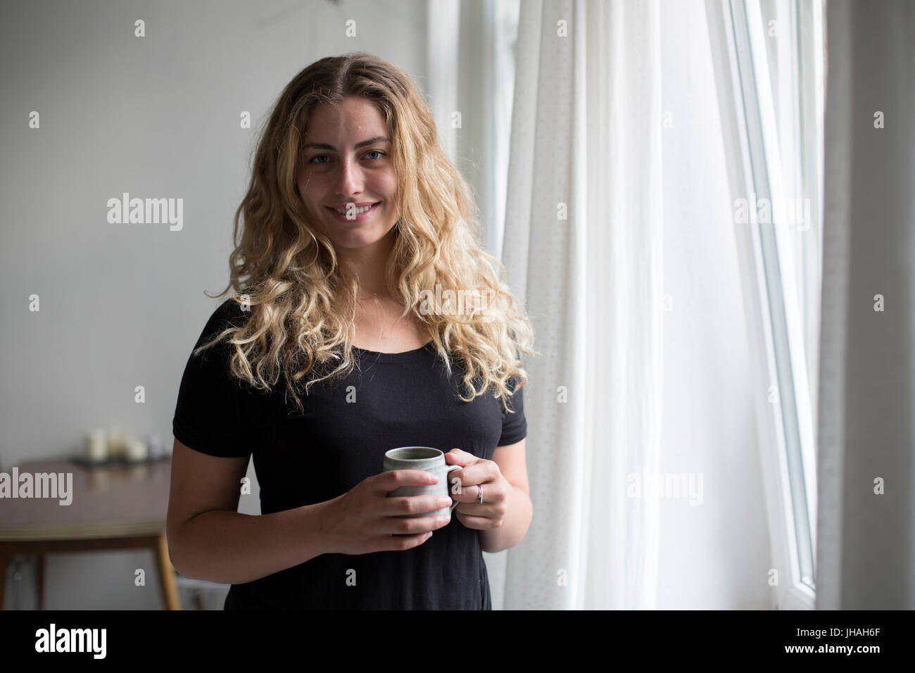 Jung, blond, schöne kaukasischen Frau bei einem heißen Getränk in der Nähe der Fenster im natürlichen Morgenlicht. Drinnen im Sommer auf einer Canon 5D aufgenommen Stockfoto