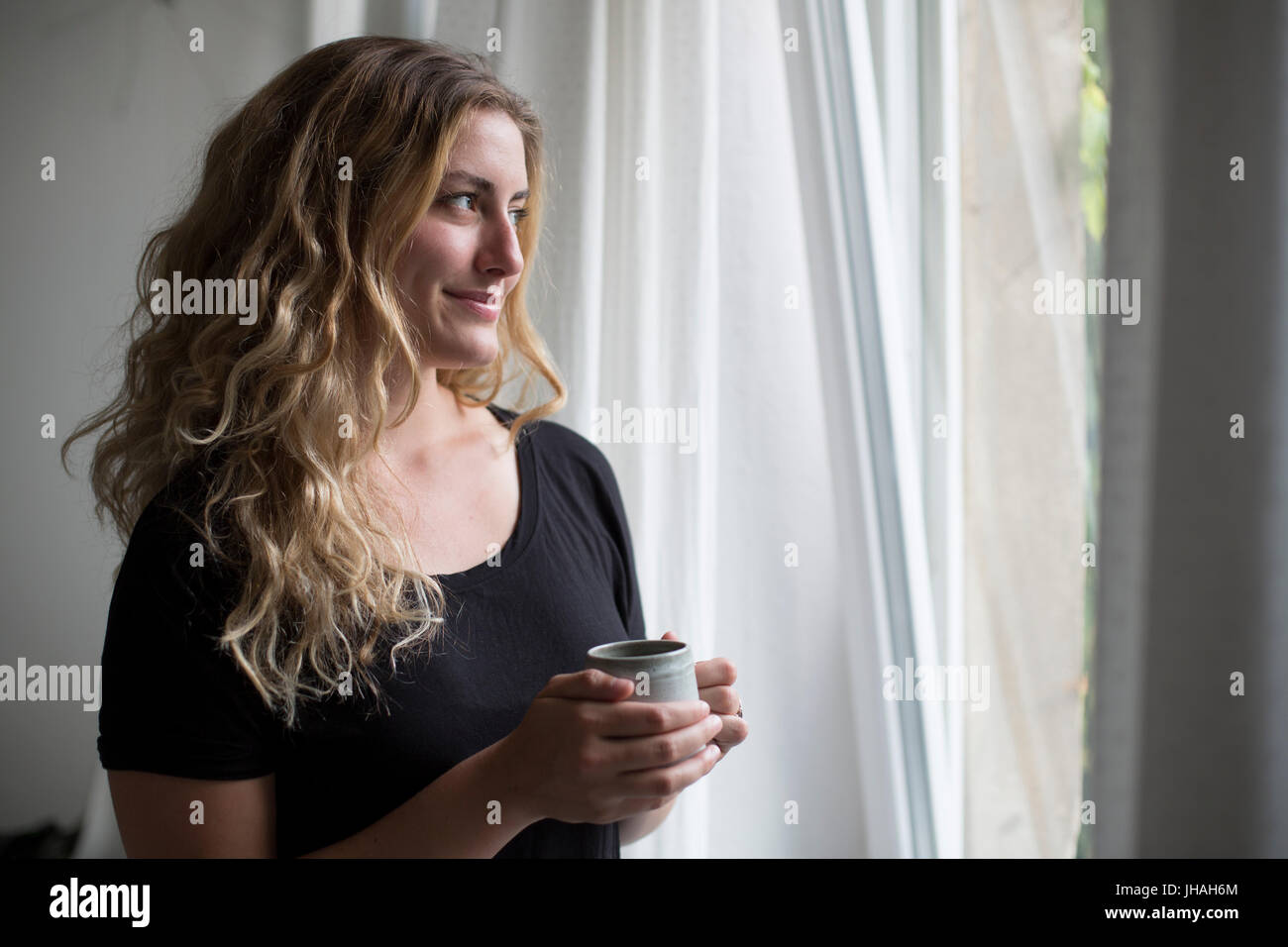 Jung, blond, schöne kaukasischen Frau bei einem heißen Getränk in der Nähe der Fenster im natürlichen Morgenlicht. Drinnen im Sommer auf einer Canon 5D aufgenommen Stockfoto