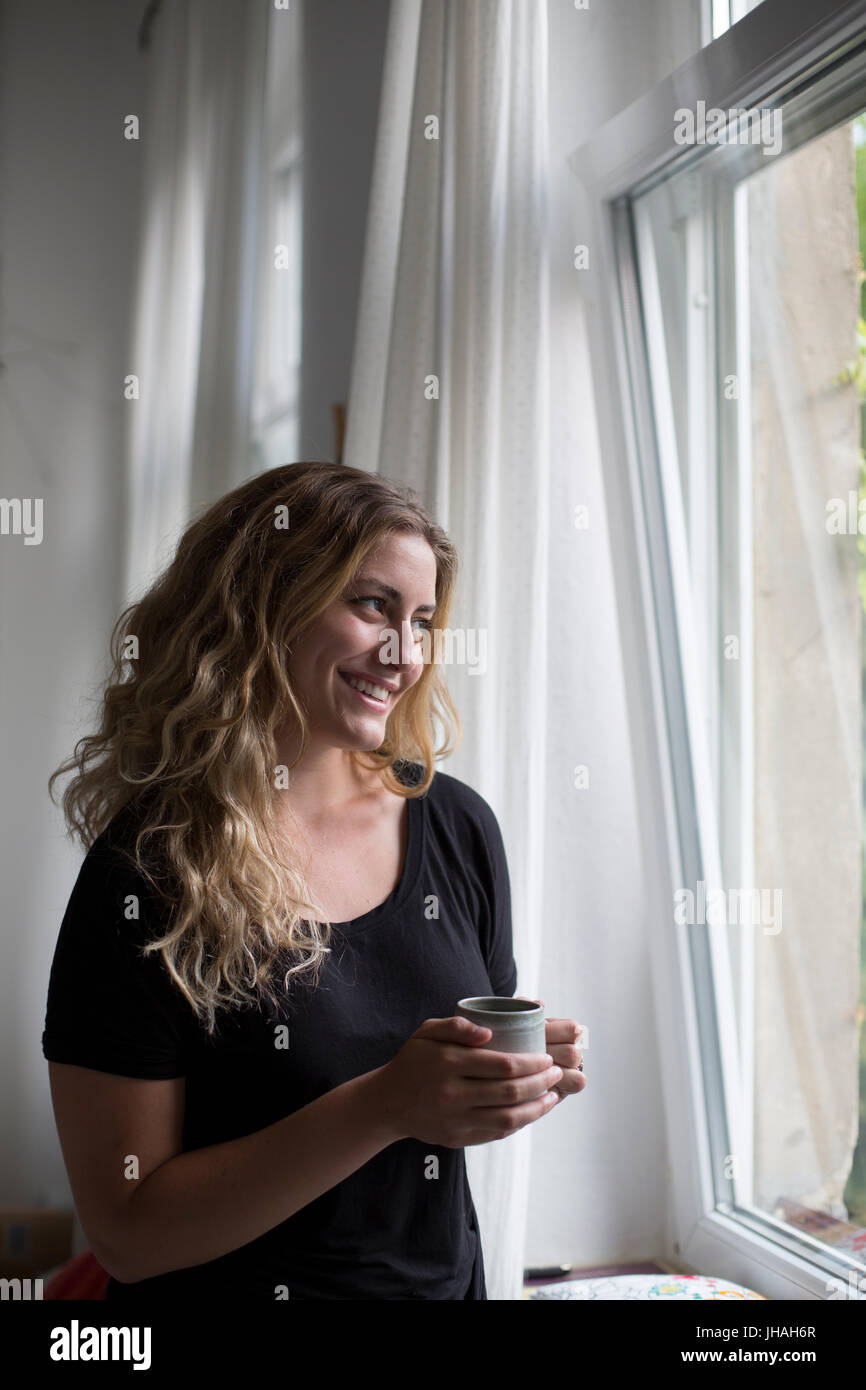 Jung, blond, schöne kaukasischen Frau bei einem heißen Getränk in der Nähe der Fenster im natürlichen Morgenlicht. Drinnen im Sommer auf einer Canon 5D aufgenommen Stockfoto