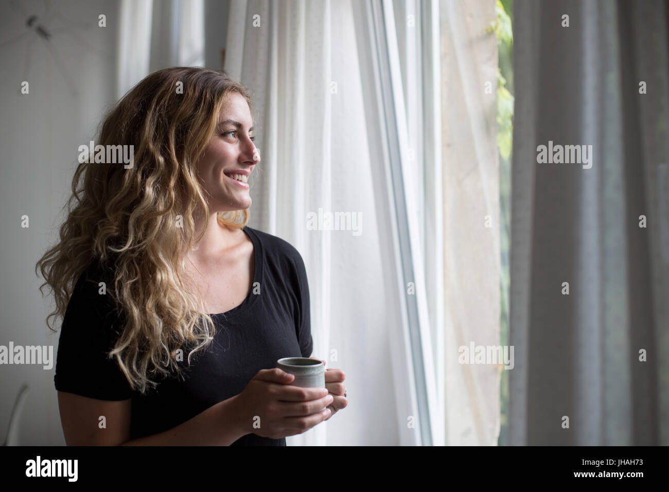 Jung, blond, schöne kaukasischen Frau bei einem heißen Getränk in der Nähe der Fenster im natürlichen Morgenlicht. Drinnen im Sommer auf einer Canon 5D aufgenommen Stockfoto