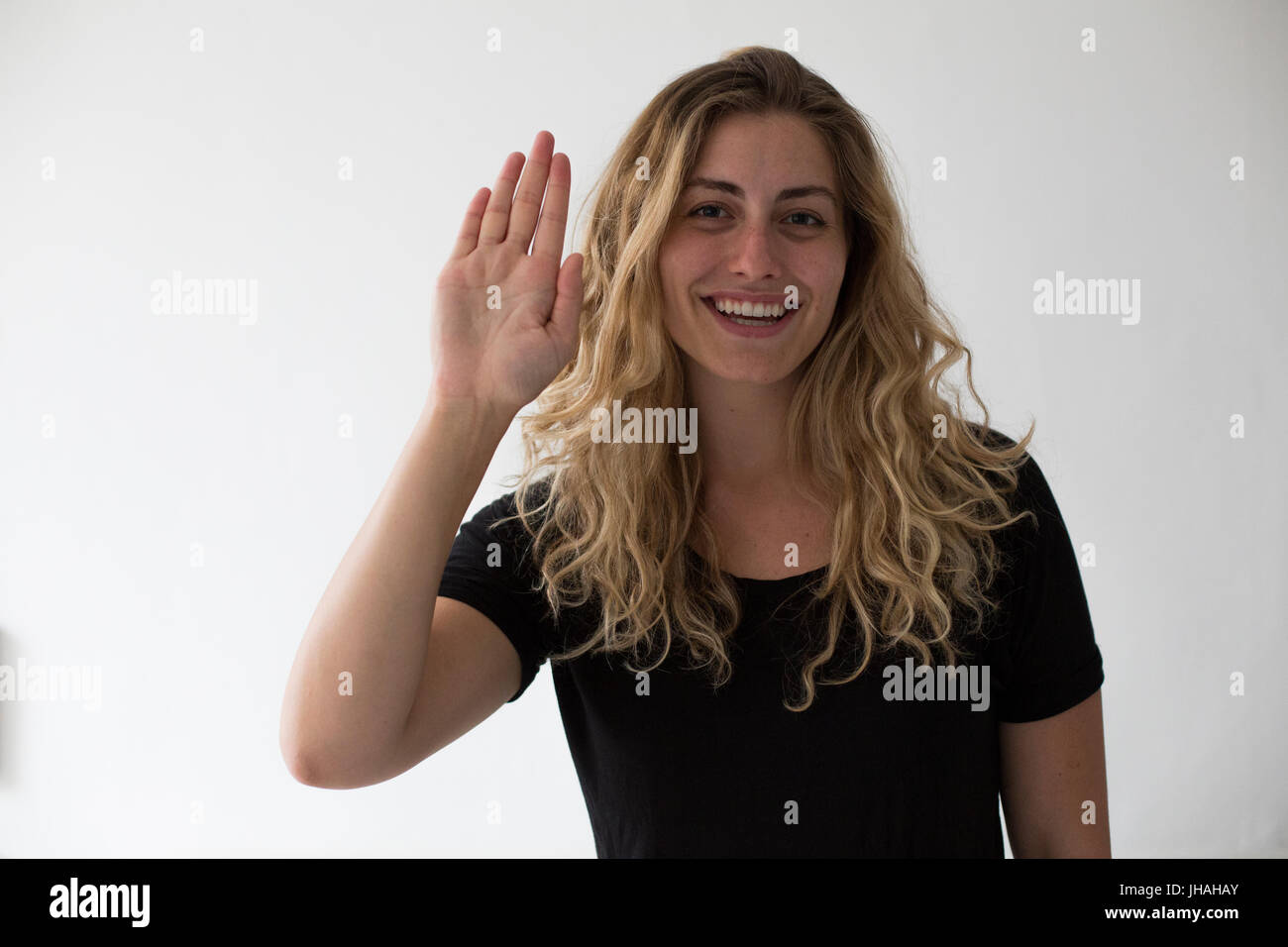 Jung, blond, kaukasischen Milennial Frau Gefühle, Freude auszudrücken und während ihre Hand auf den Betrachter vor einem weißen Hintergrund winken lächelnd. Stockfoto