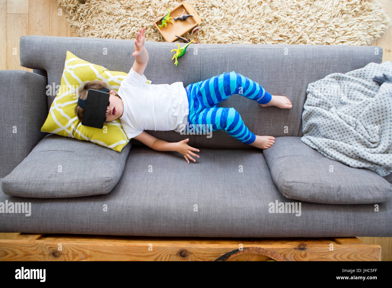 Blick von oberhalb eines kleinen Jungen auf seinem Sofa liegend, mit einem Feld von Dinosauriern. Er trägt eine virtual-Reality-Kopfhörer und hat seine Arme ausgestreckt. Stockfoto